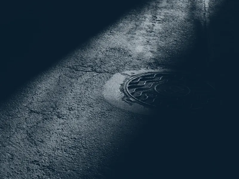 Kanaldeckel auf nassem Asphalt, von einem Lichtstreifen angeschnitten